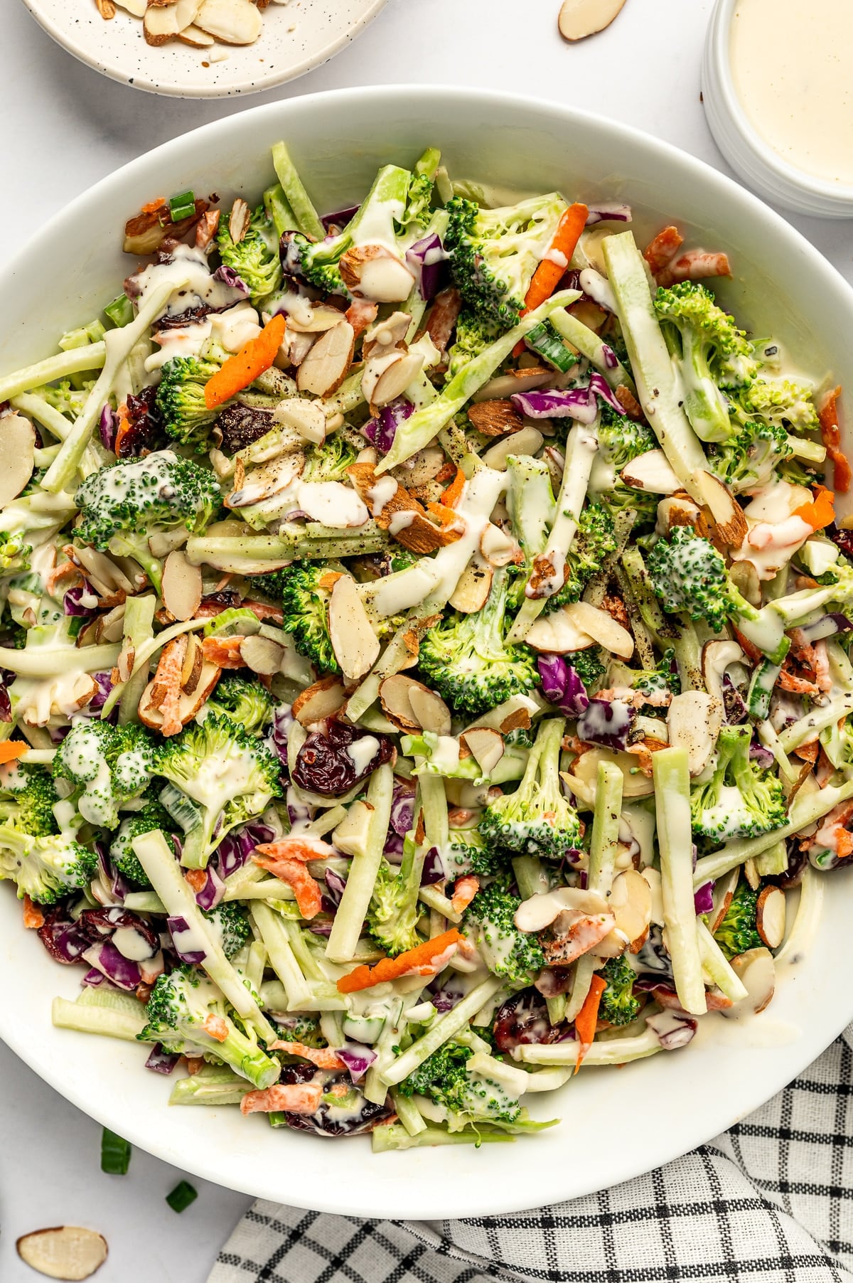 Close-up of creamy broccoli slaw in a white serving bowl with broccoli florets, broccoli stems, shredded carrots, red cabbage, dried cranberries, sliced almonds, and a drizzle of dressing.