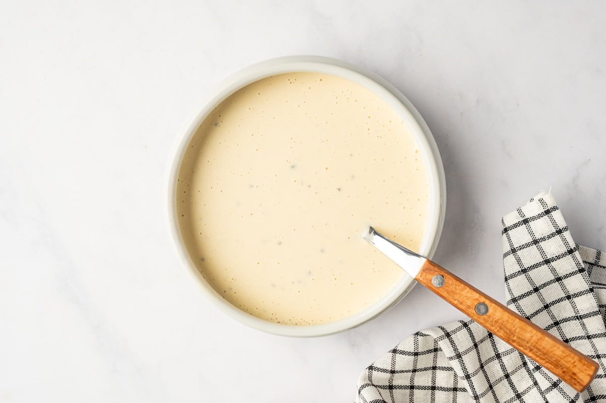Smooth creamy broccoli slaw dressing mixed in a pale oval bowl with a wooden-handled utensil, set on a white marble surface beside a black-and-white kitchen towel.