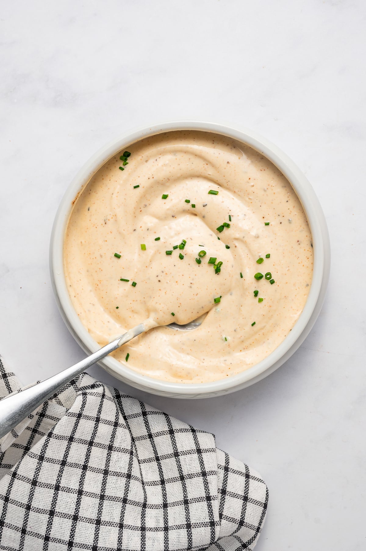 Creamy crab cake sauce mixed smoothly in a white bowl and garnished with chopped fresh chives, with a silver spoon resting in the sauce and a black-and-white checkered towel beside the bowl.
