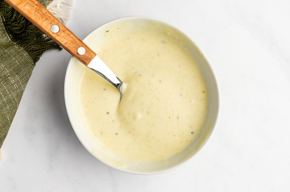 Creamy pale yellow wasabi mayo mixed in a small white bowl with a spoon, black pepper specks, and a green cloth napkin in the upper left corner.