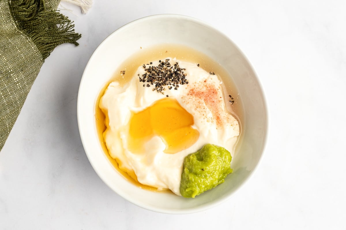 Ingredients for creamy wasabi mayo in a small white bowl, including mayonnaise, green wasabi paste, honey, salt, black pepper, and rice vinegar.