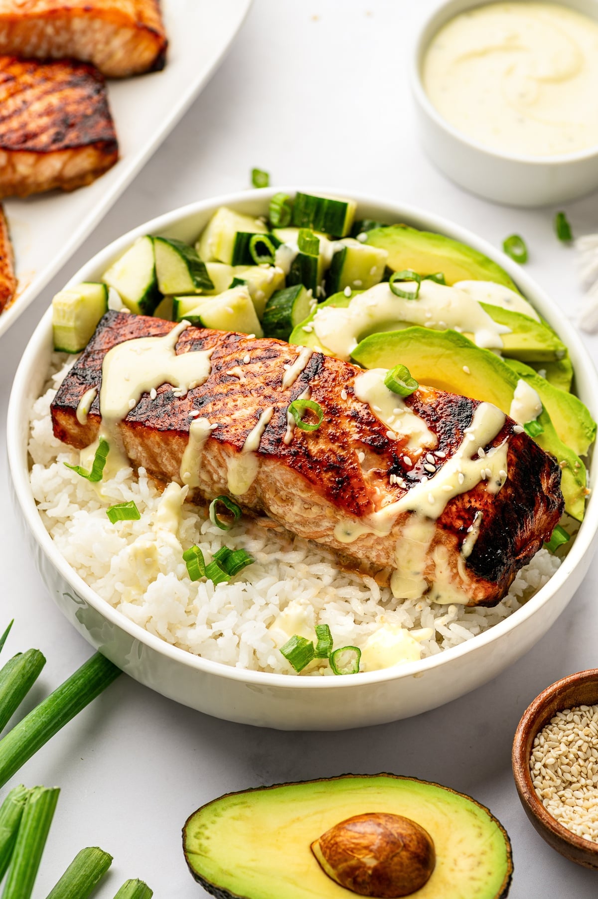 White bowl filled with fluffy rice, broiled teriyaki salmon, sliced avocado, diced cucumber, green onion, sesame seeds, and creamy wasabi mayo, with an avocado half and sesame seeds nearby.