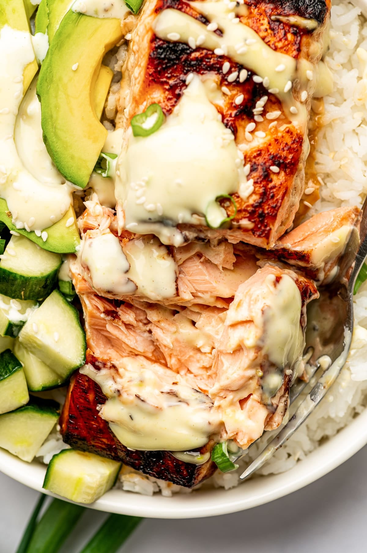 Close-up of flaky broiled teriyaki salmon being lifted with a spoon from a white rice bowl with creamy wasabi mayo, sliced avocado, diced cucumber, sesame seeds, and green onion.