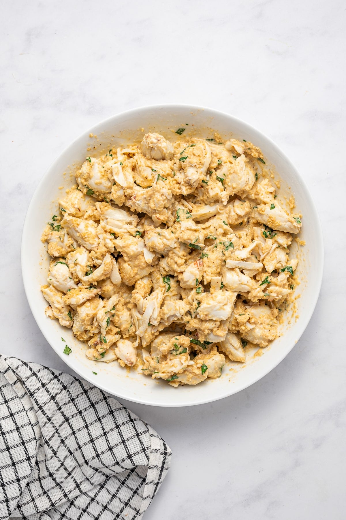 Lump crab meat gently folded with breadcrumbs and a seasoned binder in a large white bowl, with visible pieces of crab, parsley, and golden seasoning, on a white marble surface with a black-and-white checkered towel.