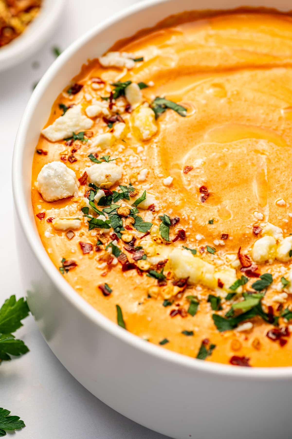 Angled close-up image of creamy, orange, Greek spicy feta dip in a white bowl, garnished with crumbled feta, chopped parsley, red pepper flakes, and a drizzle of olive oil, with fresh parsley leaves scattered across the white surface around the bowl.