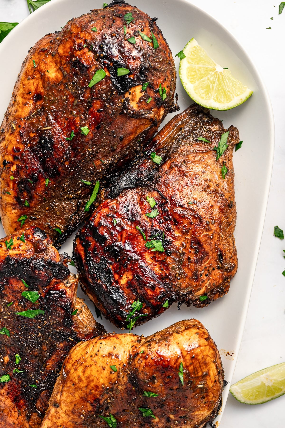Several grilled balsamic chicken breasts with dark caramelized grill marks arranged on a white oval platter, garnished with chopped parsley and served with lime wedges on a white marble surface.