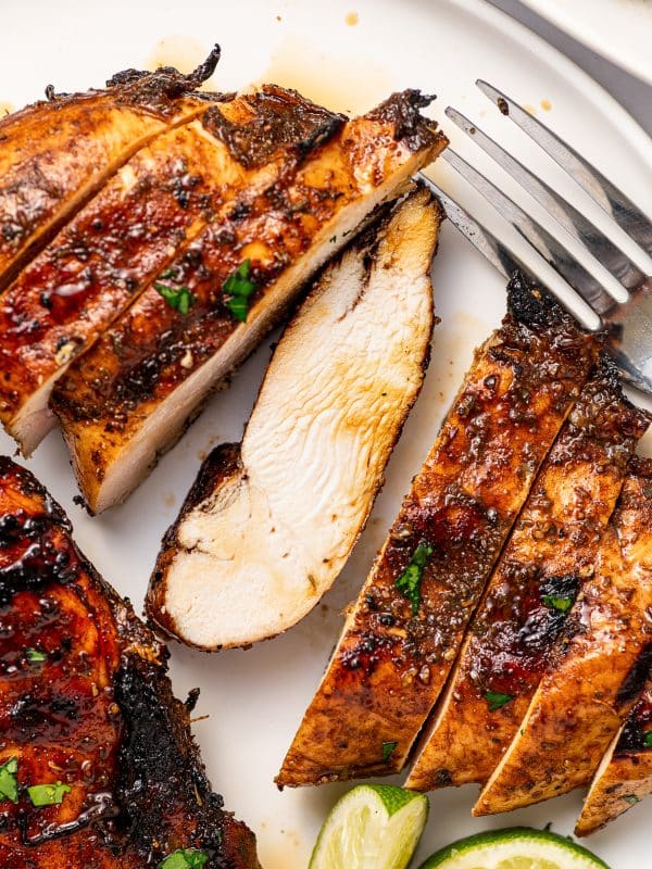 Close-up of sliced grilled chicken breast with a browned balsamic exterior and pale juicy center on a white plate, beside a silver fork, sliced grilled chicken, chopped parsley, and lime wedges.