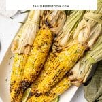 Pinterest image showing grilled corn on the cob with charred yellow kernels and tied-back pale green husks served on a white oval platter with lime wedges, chopped green herbs, flaky salt, butter in a white dish, and a green cloth napkin. Black text overlay in a white box reads "HOW TO GRILL CORN ON THE COB" and "THEFORKEDSPOON.COM" with a thin beige line.