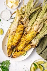 Grilled corn on the cob with husks tied back served on a white oval platter with lime wedges, chopped green herbs, flaky salt, butter in a small white dish, and a green cloth napkin.
