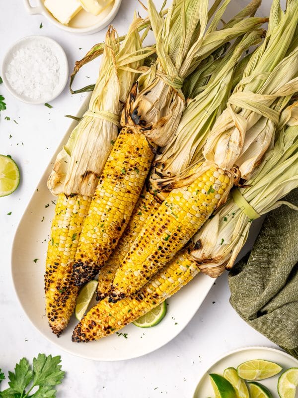 Grilled corn on the cob with husks tied back served on a white oval platter with lime wedges, chopped green herbs, flaky salt, butter in a small white dish, and a green cloth napkin.
