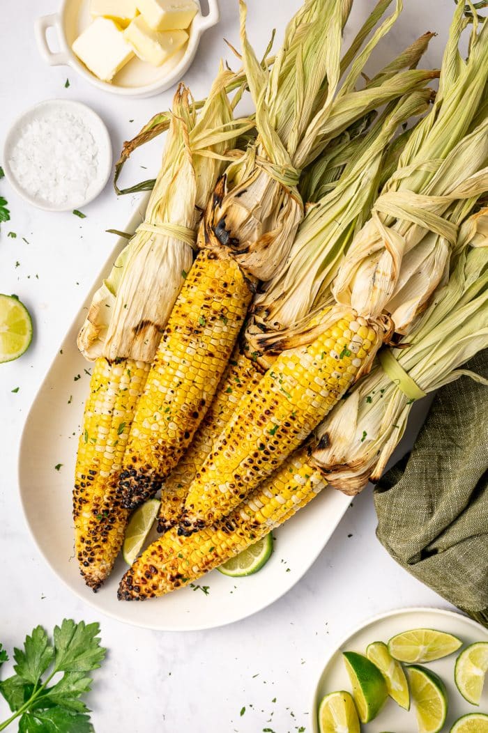 Grilled corn on the cob with husks tied back served on a white oval platter with lime wedges, chopped green herbs, flaky salt, butter in a small white dish, and a green cloth napkin.