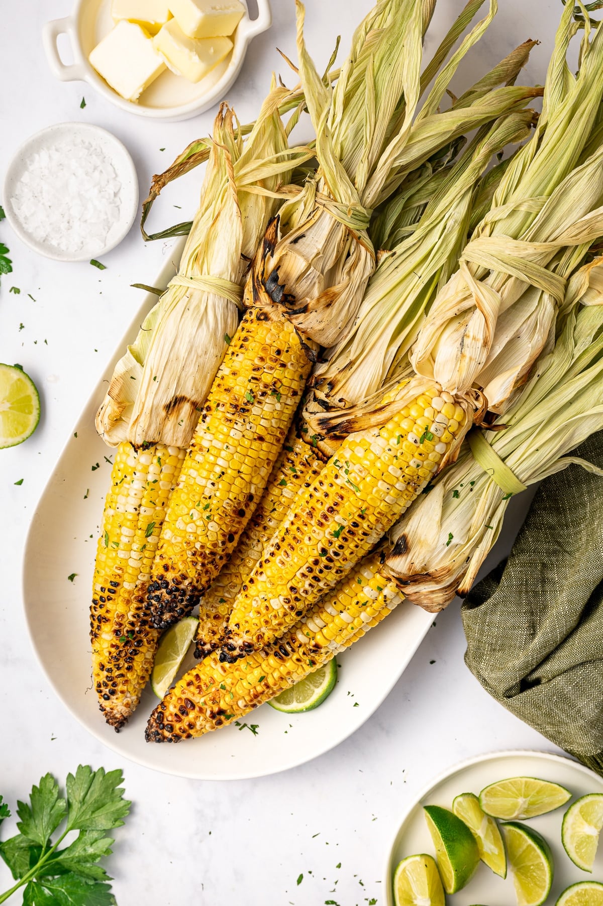 Grilled corn on the cob with husks tied back served on a white oval platter with lime wedges, chopped green herbs, flaky salt, butter in a small white dish, and a green cloth napkin.