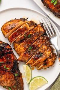 Overhead view of sliced grilled chicken breast with a browned balsamic exterior and light juicy interior on a white plate, surrounded by grilled chicken, lime wedges, parsley, and a silver fork with a wooden handle.
