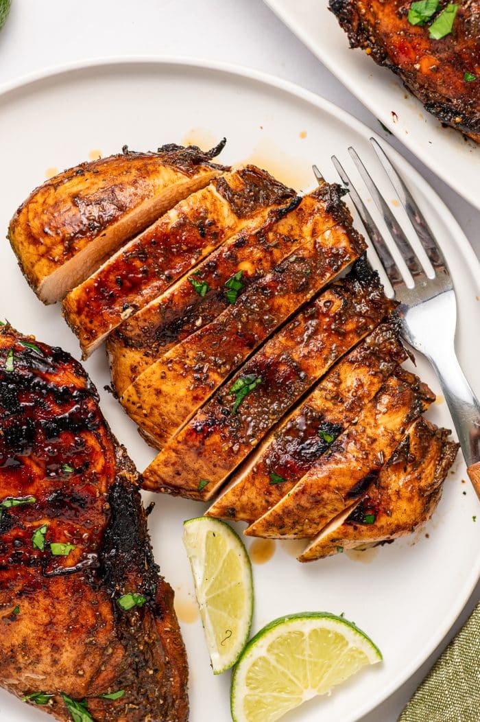 Overhead view of sliced grilled chicken breast with a browned balsamic exterior and light juicy interior on a white plate, surrounded by grilled chicken, lime wedges, parsley, and a silver fork with a wooden handle.