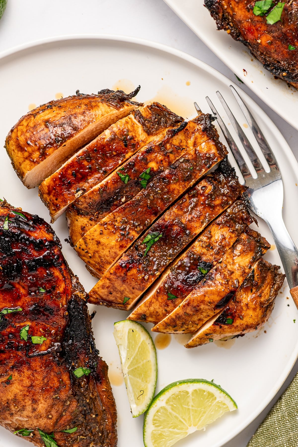 Overhead view of sliced grilled chicken breast with a browned balsamic exterior and light juicy interior on a white plate, surrounded by grilled chicken, lime wedges, parsley, and a silver fork with a wooden handle.