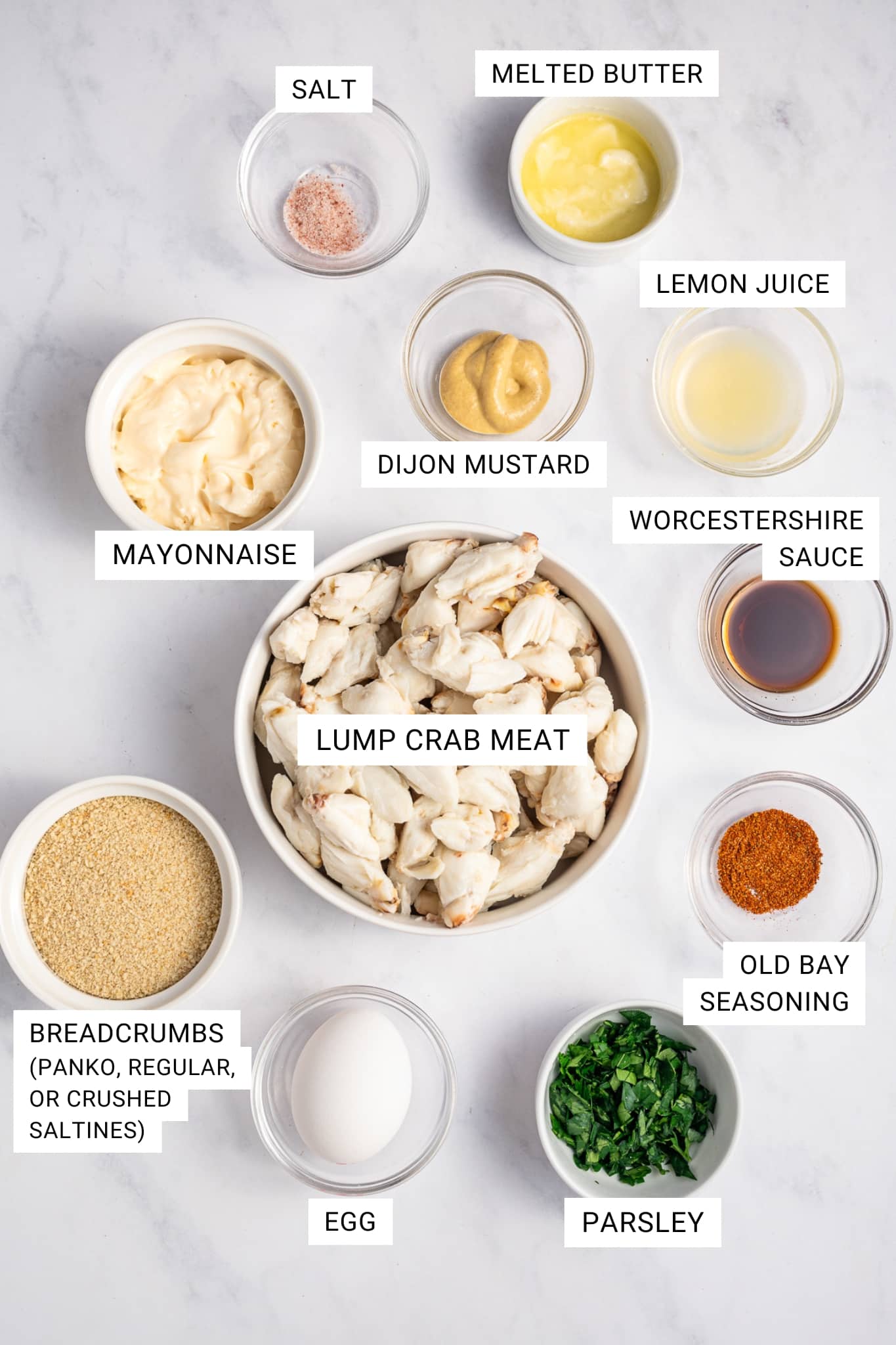 Labeled ingredient image for crab cakes showing mayonnaise, breadcrumbs labeled "Breadcrumbs (panko, regular, or crushed saltines)," egg, parsley, Old Bay seasoning, Worcestershire sauce, lump crab meat, Dijon mustard, lemon juice, melted butter, and salt arranged in small bowls on a white marble surface. Text labels identify each ingredient.