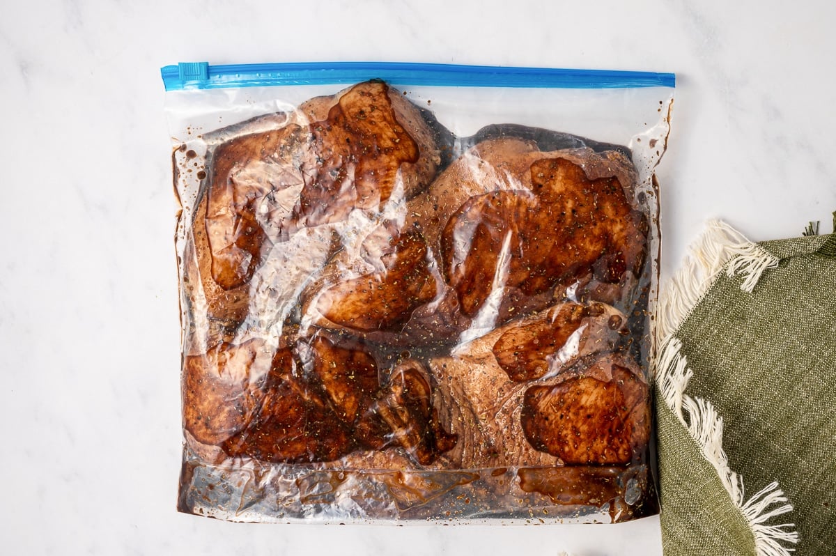 A sealed clear zip-top bag filled with raw chicken breasts coated in dark balsamic marinade, placed on a white marble surface beside an olive green fringed kitchen towel.