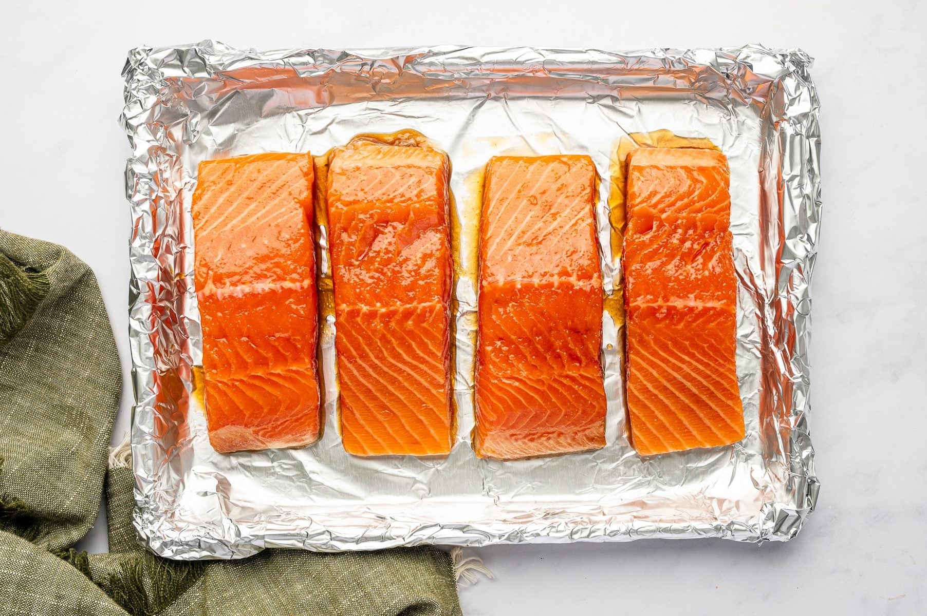 Four raw marinated salmon fillets arranged skin-side down on a foil-lined baking sheet, ready to be broiled, with a green cloth napkin along the right and bottom edges.