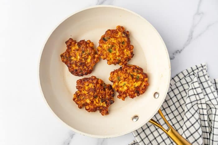 Four golden brown corn fritters cooking in a cream-colored skillet with crisp edges and visible corn, green onion, and jalapeño pieces.