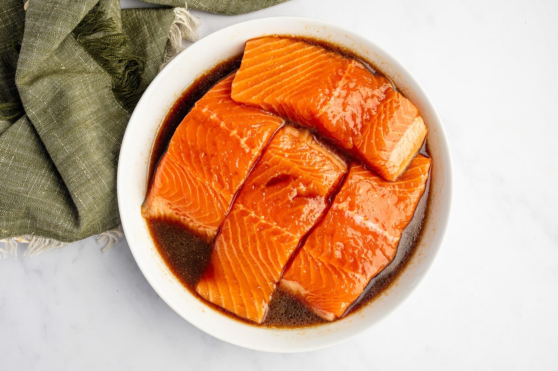 Raw salmon fillets marinating in a white shallow bowl filled with dark teriyaki sauce, shown from above with a green cloth napkin tucked along the upper left side.