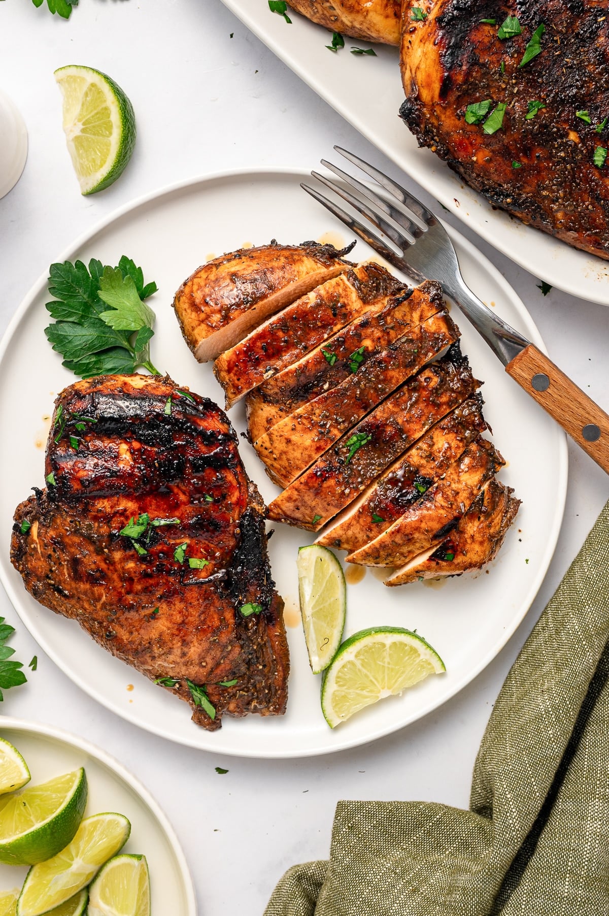 Sliced grilled chicken breast with dark balsamic grill marks arranged on a white plate beside a whole grilled chicken breast, lime wedges, chopped parsley, and a silver fork with a wooden handle.