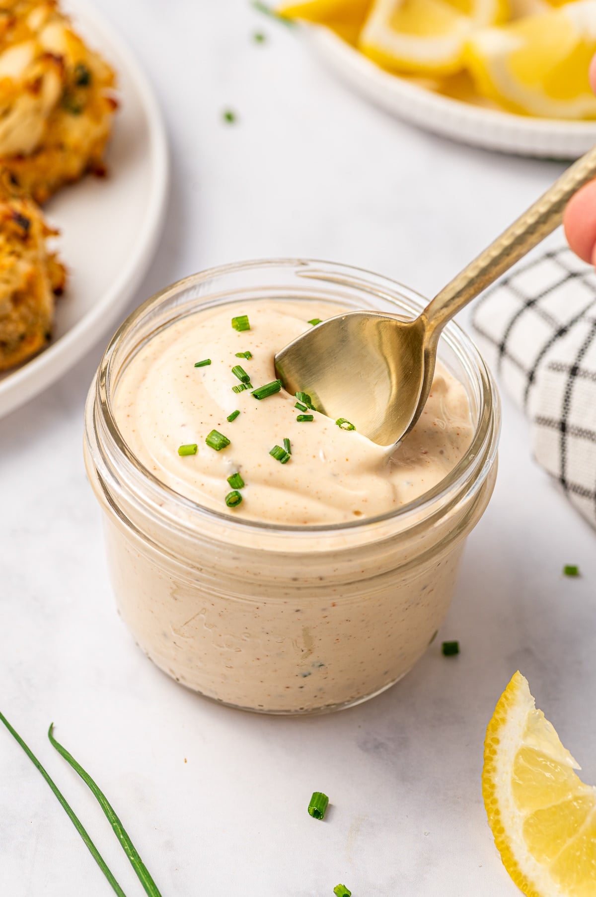 A gold spoon dipping into creamy crab cake sauce in a clear glass jar, topped with chopped chives and served with lemon wedges, crab cakes, and a black-and-white checkered towel.