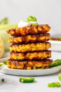 Stack of crispy corn fritters on a white plate topped with sour cream and green onion, with sliced jalapeños and lime in the background.