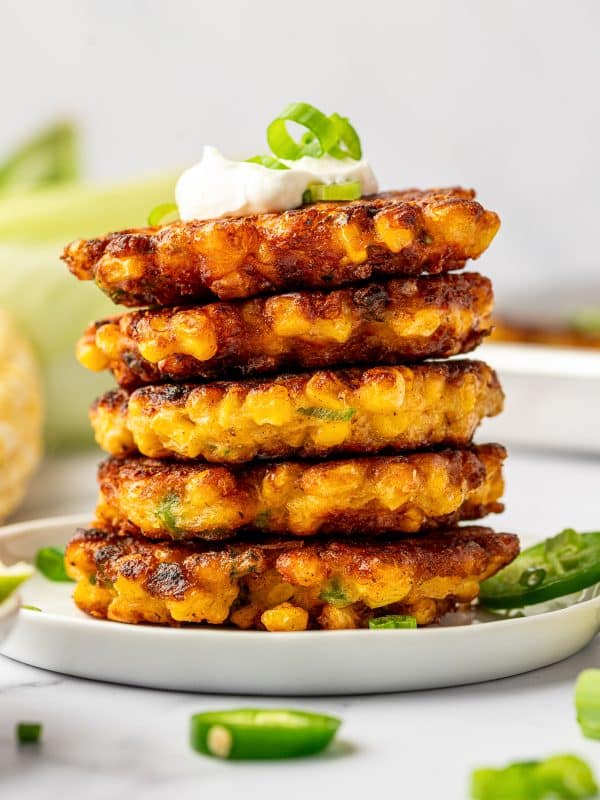 Stack of crispy corn fritters on a white plate topped with sour cream and green onion, with sliced jalapeรฑos and lime in the background.
