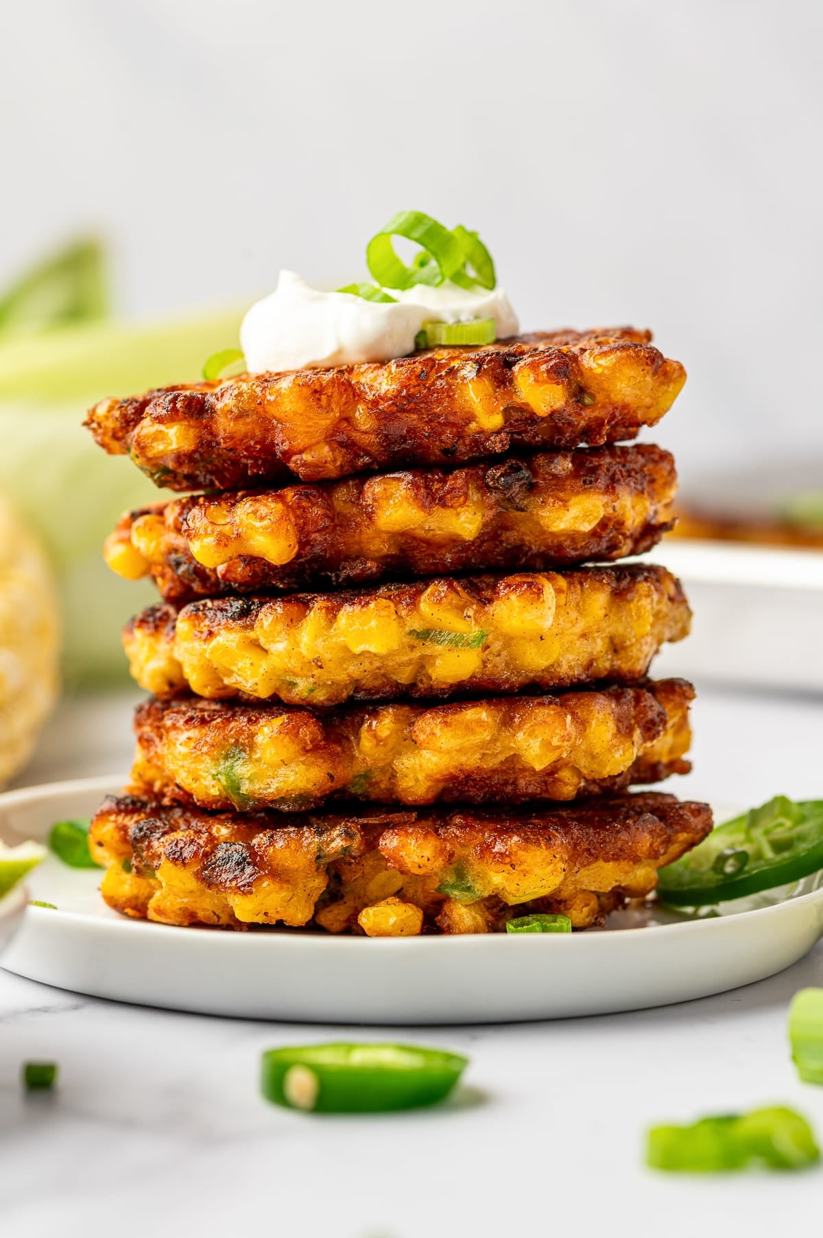 Stack of crispy corn fritters on a white plate topped with sour cream and green onion, with sliced jalapeños and lime in the background.
