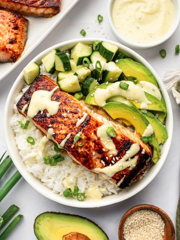 Teriyaki salmon rice bowl served in a white bowl with fluffy white rice, broiled salmon, diced cucumber, sliced avocado, green onion, sesame seeds, and creamy wasabi mayo drizzled over the top.