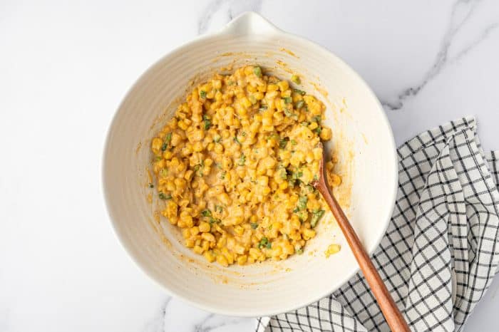 Thick corn fritter batter with corn, cheddar cheese, green onions, and jalapeños mixed in a cream-colored bowl with a wooden spoon.