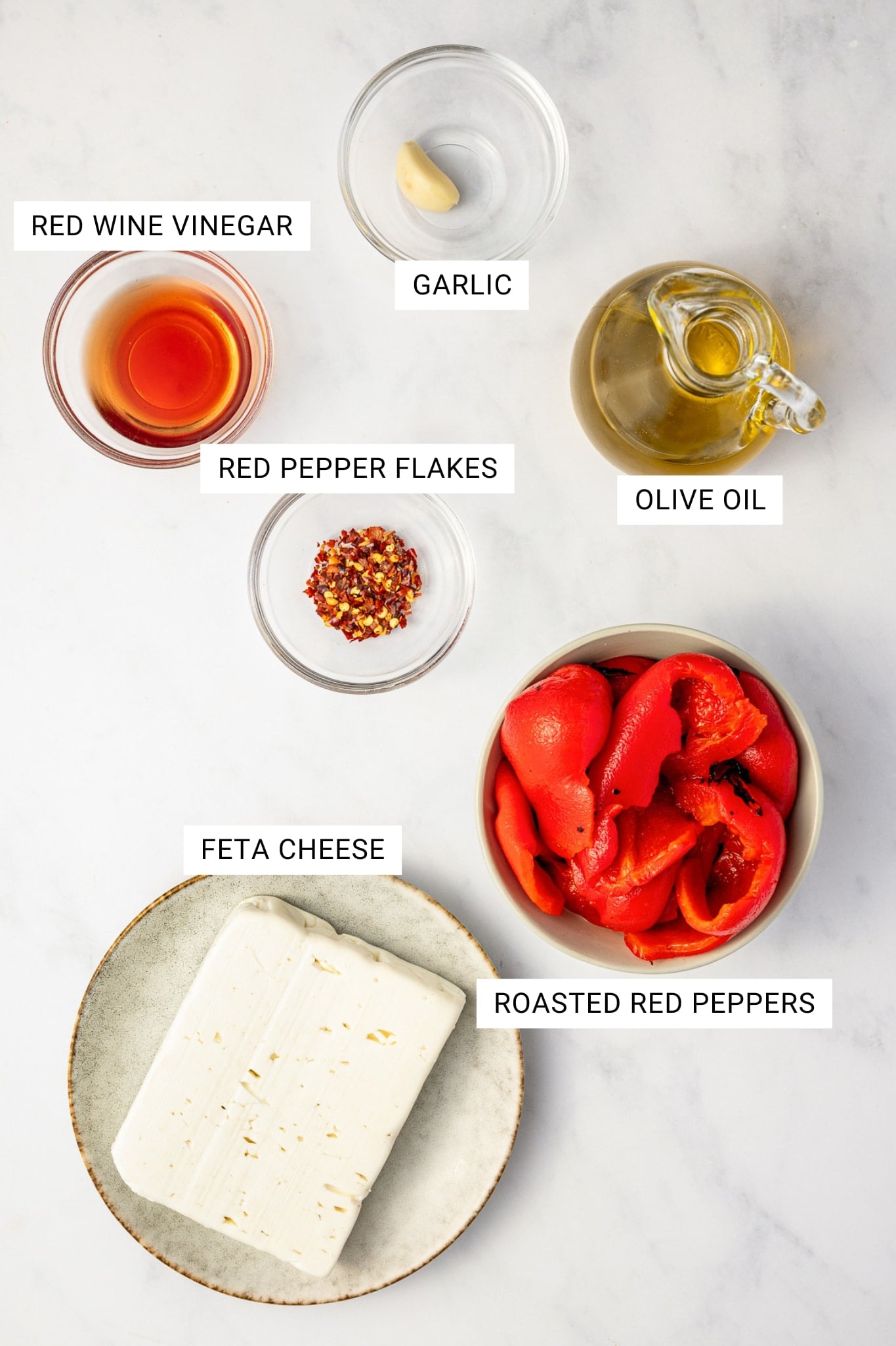 Overhead ingredient image for tirokafteri showing a block of feta cheese on a cream plate, roasted red peppers in a bowl, olive oil in a glass cruet, red wine vinegar in a small glass bowl, red pepper flakes in a small glass bowl, and a garlic clove in a small glass bowl, with white text labels for each ingredient.