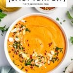 Pinterest pin image of creamy orange tirokafteri in a white bowl topped with crumbled feta, red pepper flakes, chopped parsley, and olive oil, with a glass bottle of olive oil, red pepper flakes, Kalamata olives, parsley, and a pale blue napkin in the background. A white text box reads "TIROKAFTERI (SPICY FETA DIP)" and "THEFORKEDSPOON.COM" with a green line divider.