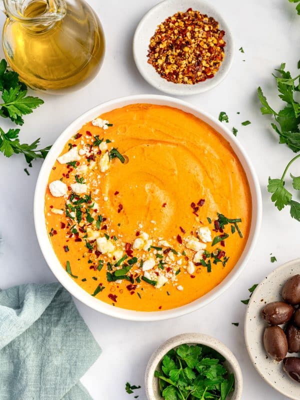 Overhead image of creamy orange tirokafteri in a white bowl topped with crumbled feta, red pepper flakes, chopped parsley, and olive oil, with a glass bottle of olive oil, a small white bowl of red pepper flakes, Kalamata olives, fresh parsley, and a pale blue napkin on a white marble surface.