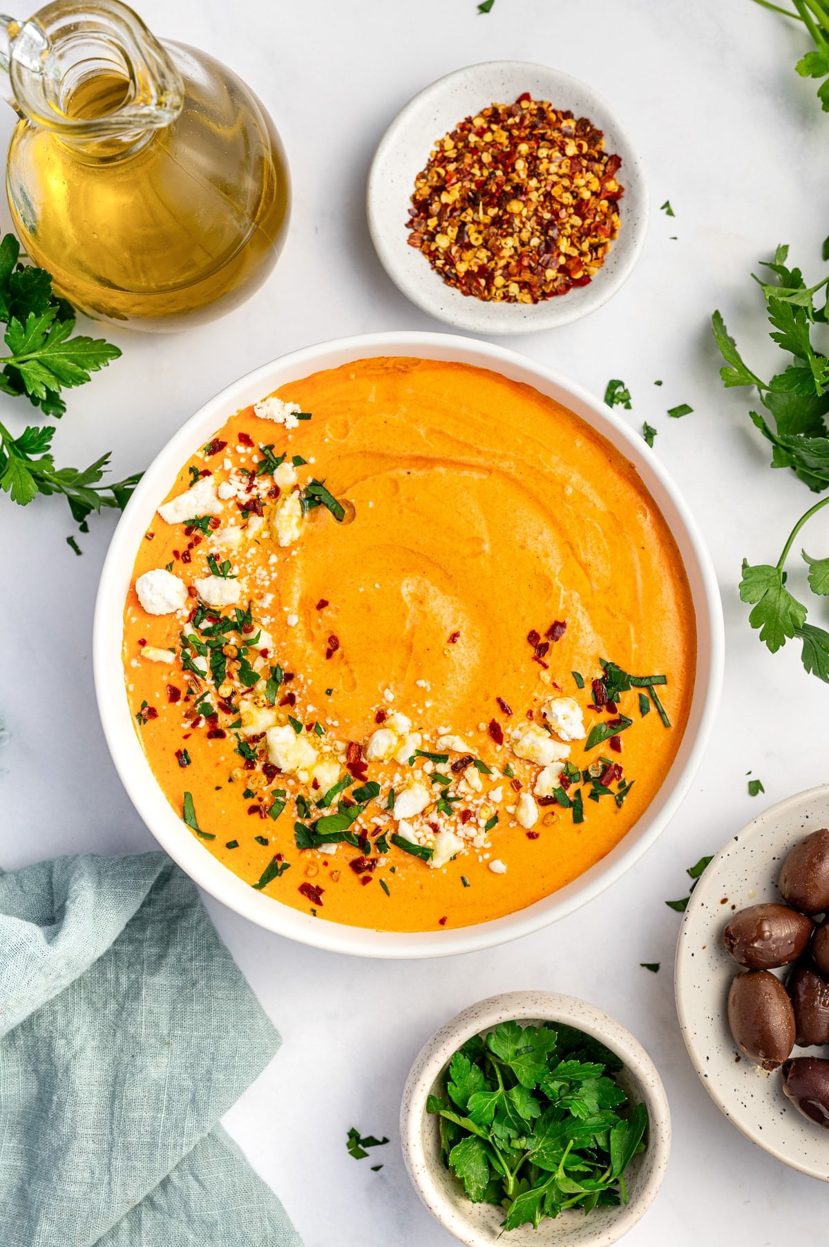 Overhead image of creamy orange tirokafteri in a white bowl topped with crumbled feta, red pepper flakes, chopped parsley, and olive oil, with a glass bottle of olive oil, a small white bowl of red pepper flakes, Kalamata olives, fresh parsley, and a pale blue napkin on a white marble surface.