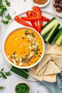 White bowl of creamy orange tirokafteri topped with crumbled feta, red pepper flakes, and chopped parsley, served on a white platter with cucumber spears, red bell pepper strips, pita bread triangles, Kalamata olives, fresh parsley, and a pale blue napkin.