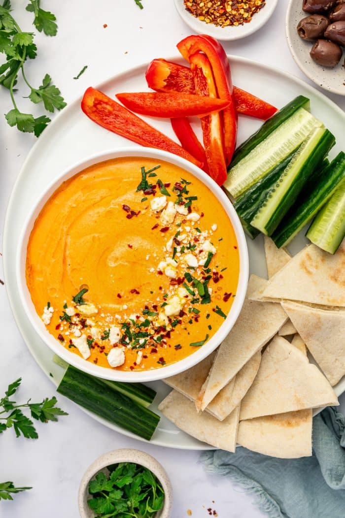 White bowl of creamy orange tirokafteri topped with crumbled feta, red pepper flakes, and chopped parsley, served on a white platter with cucumber spears, red bell pepper strips, pita bread triangles, Kalamata olives, fresh parsley, and a pale blue napkin.