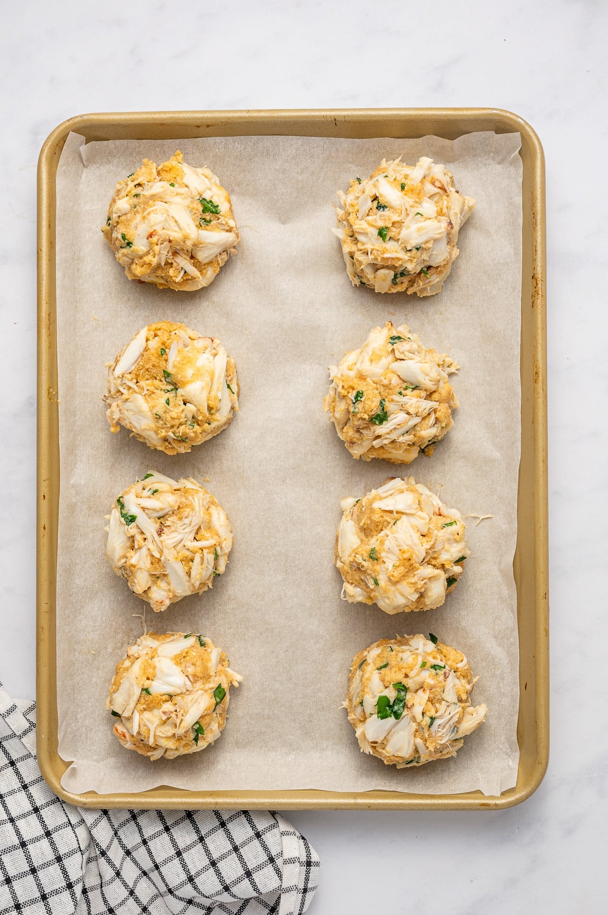 Eight mounded, unbaked crab cakes made with visible lump crab meat and parsley are arranged on a parchment-lined gold baking sheet beside a black and white checkered towel.