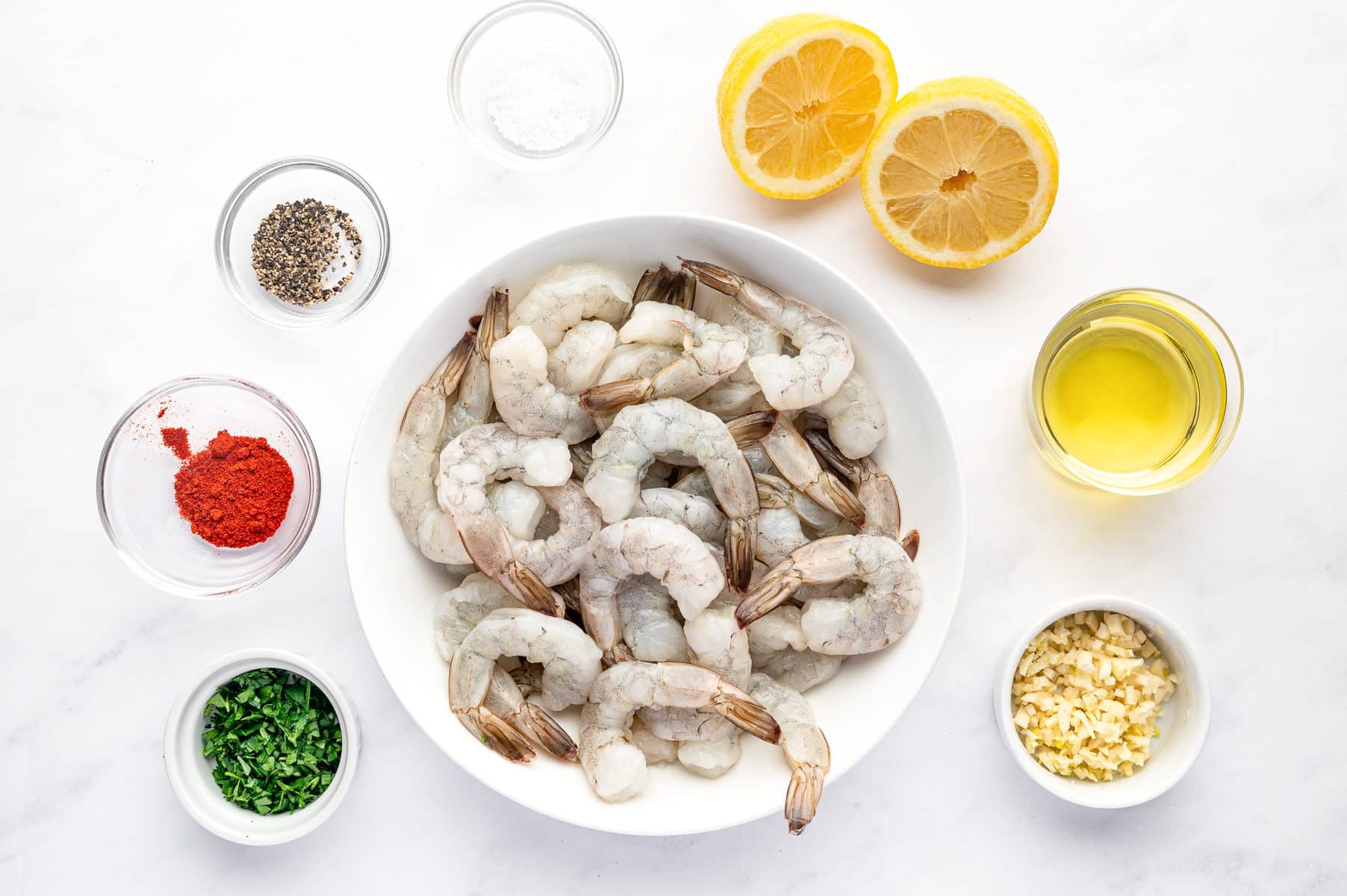 Overhead ingredient layout on a white background showing a white plate of raw large shrimp surrounded by olive oil, minced garlic, halved lemons, kosher salt, black pepper, paprika, and minced parsley.