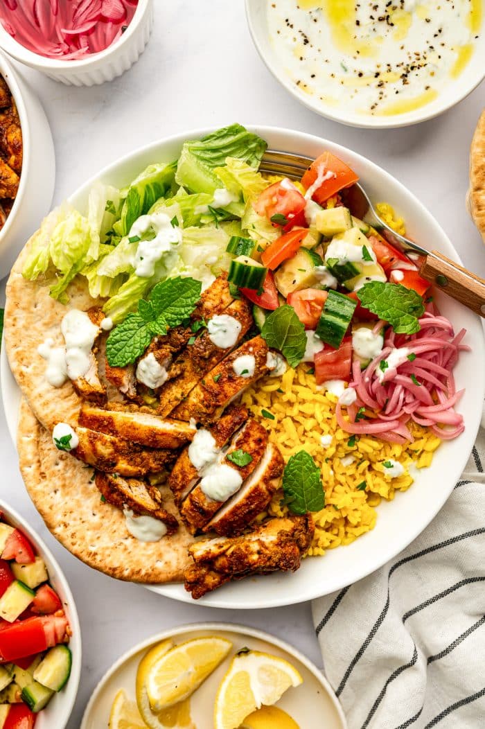 A large white chicken shawarma bowl filled with golden yellow rice, sliced spiced chicken, chopped romaine, cucumber tomato salad, pink pickled red onions, white tzatziki, mint, parsley, and pita, with lemon wedges and side dishes surrounding the bowl.