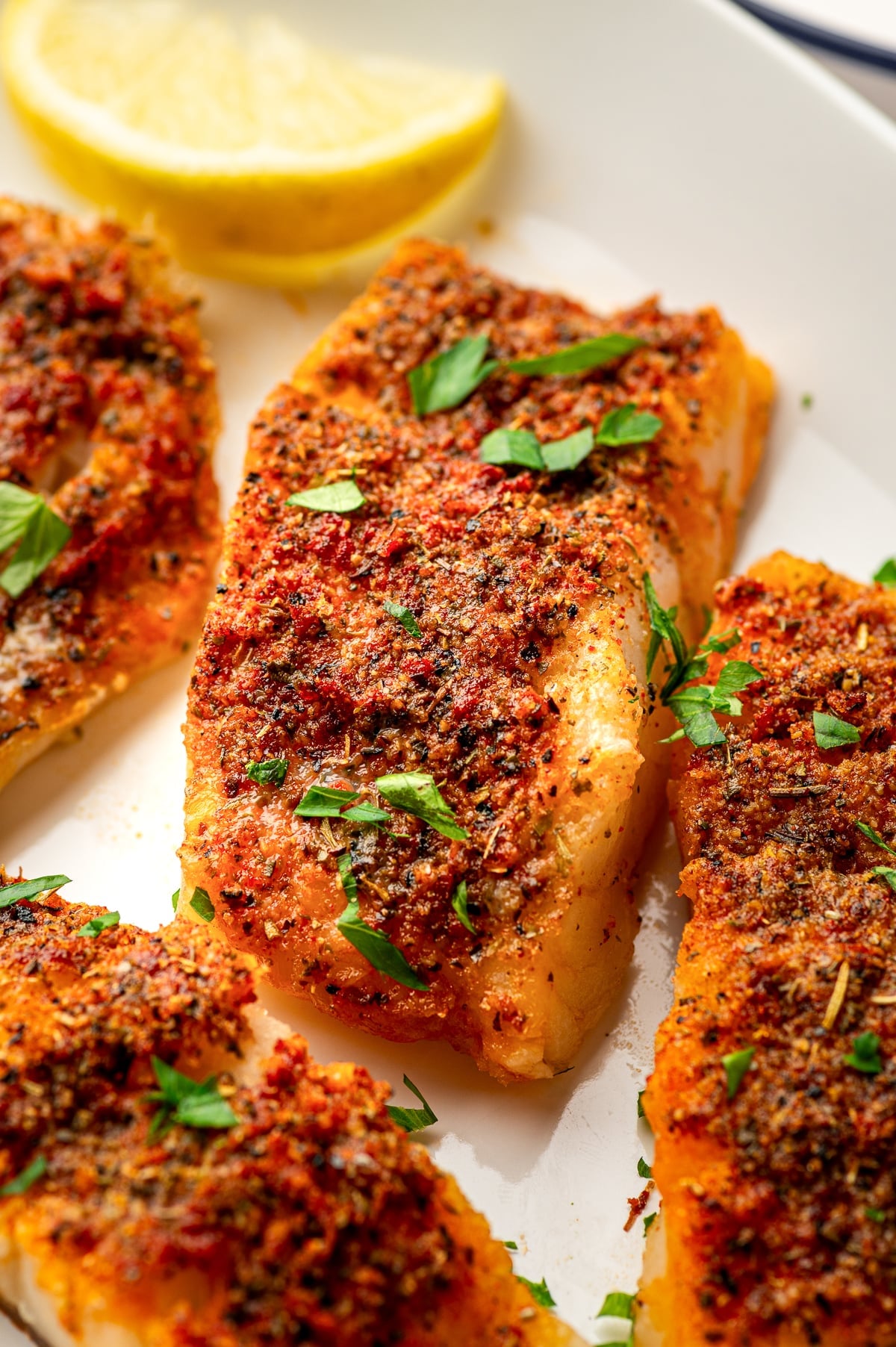 Close-up of a thick golden baked cod fillet coated with paprika, dried herbs, black pepper, and lemon zest on a cream-colored plate, garnished with chopped parsley. A lemon wedge and additional cod fillets appear softly blurred in the background.