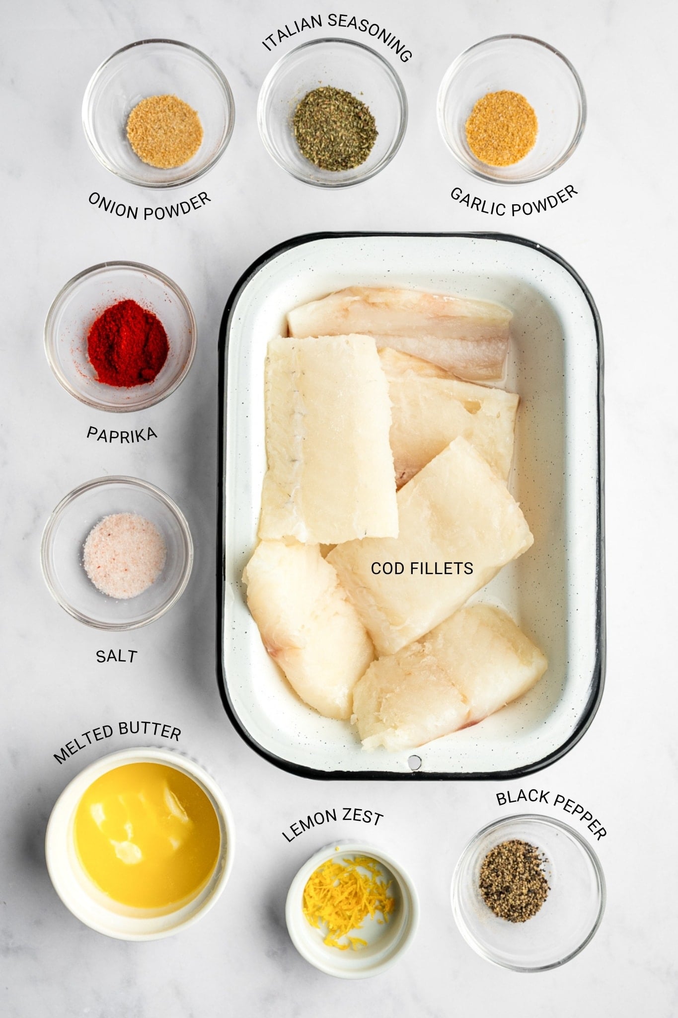Labeled overhead ingredient photo with raw cod fillets in a white enamel baking dish with a black rim, surrounded by small clear bowls of paprika, salt, black pepper, garlic powder, Italian seasoning, and onion seasoning, plus white bowls of melted yellow butter and lemon zest. Black text labels identify each ingredient.
