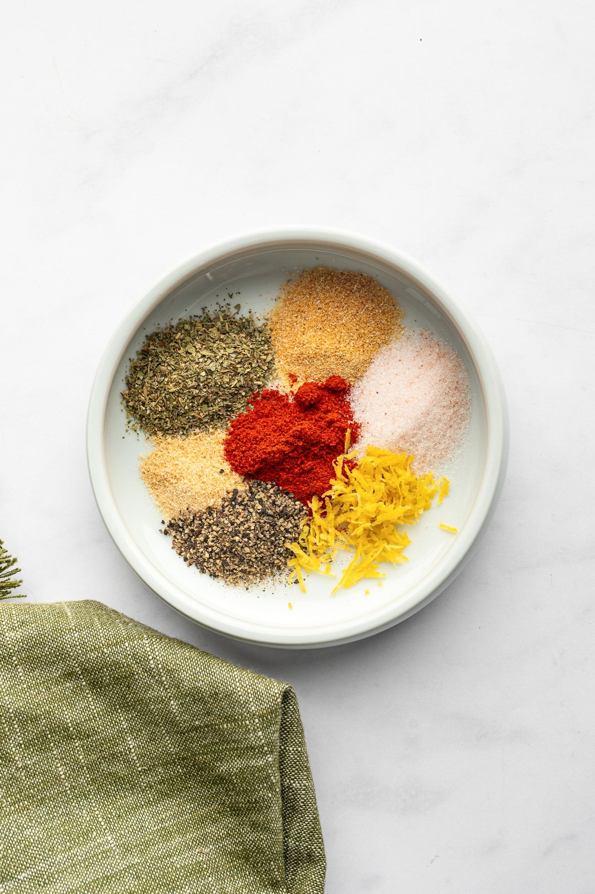Separate piles of Italian seasoning, garlic powder, onion powder, paprika, pink salt, black pepper, and fresh lemon zest arranged in a shallow pale gray ceramic bowl beside an olive-green cloth napkin.