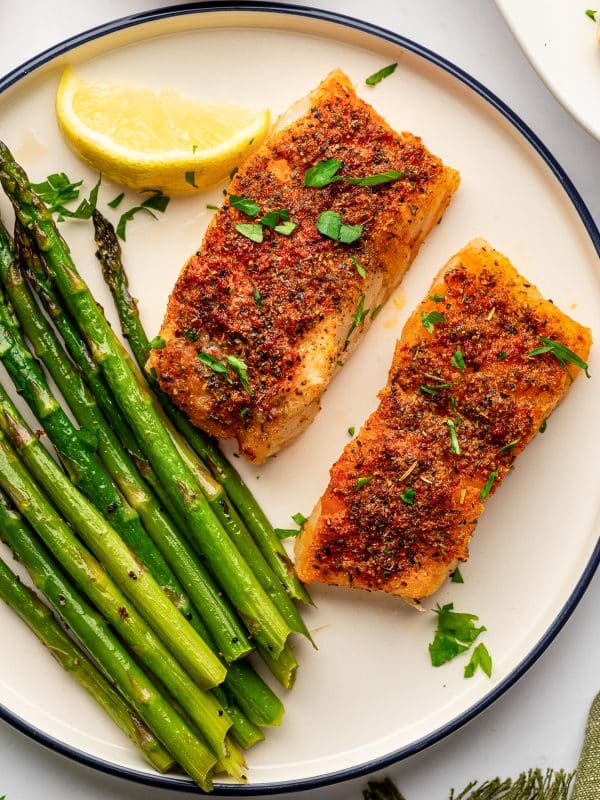Two golden baked cod fillets served with roasted green asparagus on a cream-colored round plate with a dark navy rim. The fish is garnished with chopped parsley and served with a lemon wedge, with a green fringed napkin at the lower right.