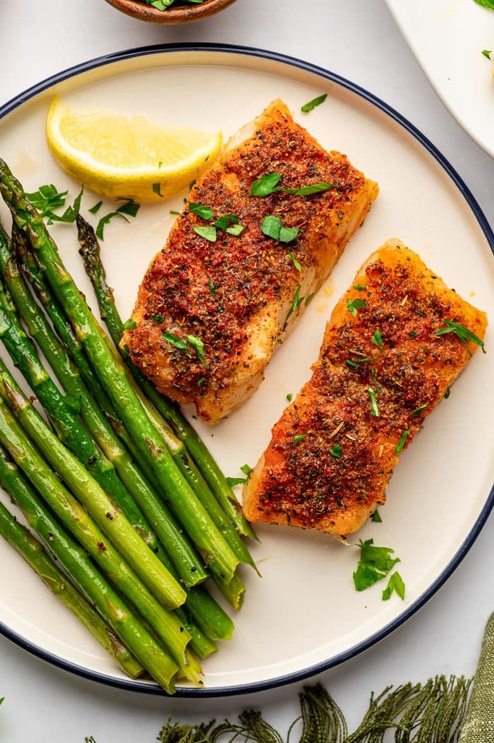 Two golden baked cod fillets served with roasted green asparagus on a cream-colored round plate with a dark navy rim. The fish is garnished with chopped parsley and served with a lemon wedge, with a green fringed napkin at the lower right.