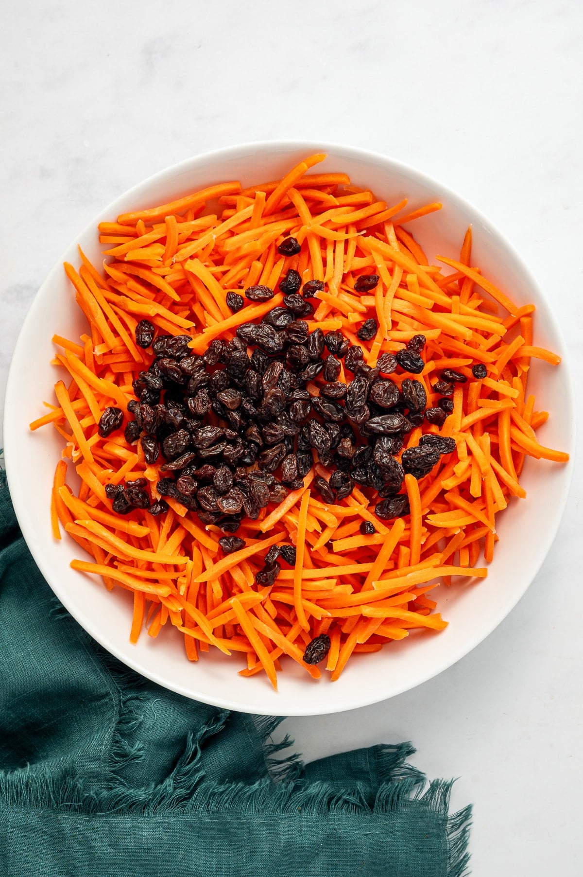 Shredded orange carrots topped with dark raisins in a white oval bowl on a white marble surface with a dark green cloth napkin tucked underneath.