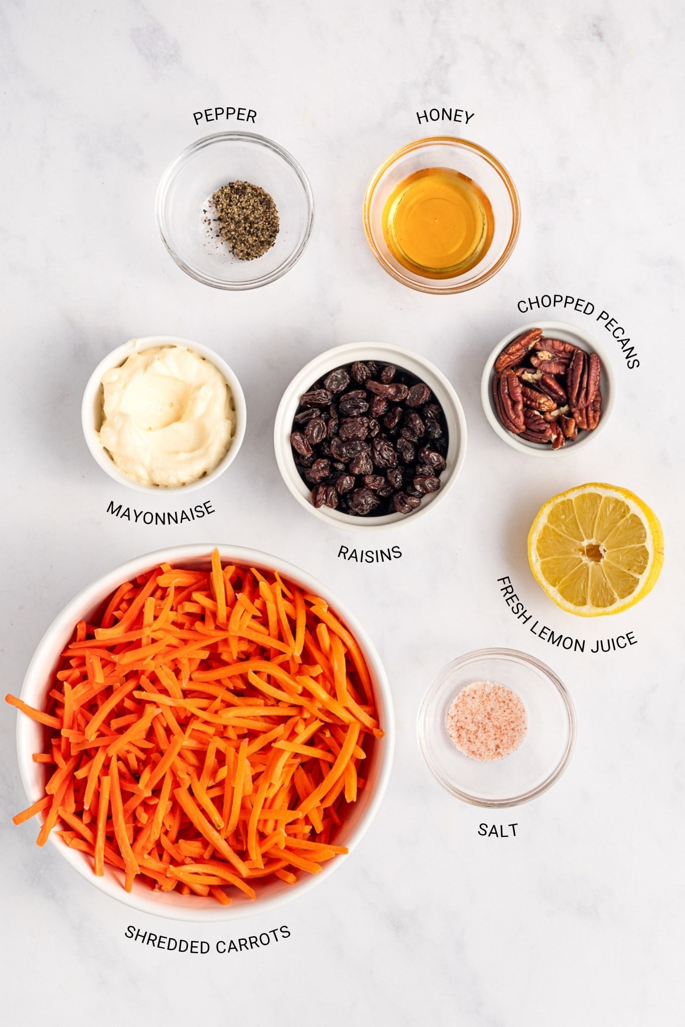 Labeled ingredients for carrot raisin salad on a white marble surface, including shredded carrots, raisins, mayonnaise, honey, fresh lemon juice, salt, pepper, and chopped pecans.