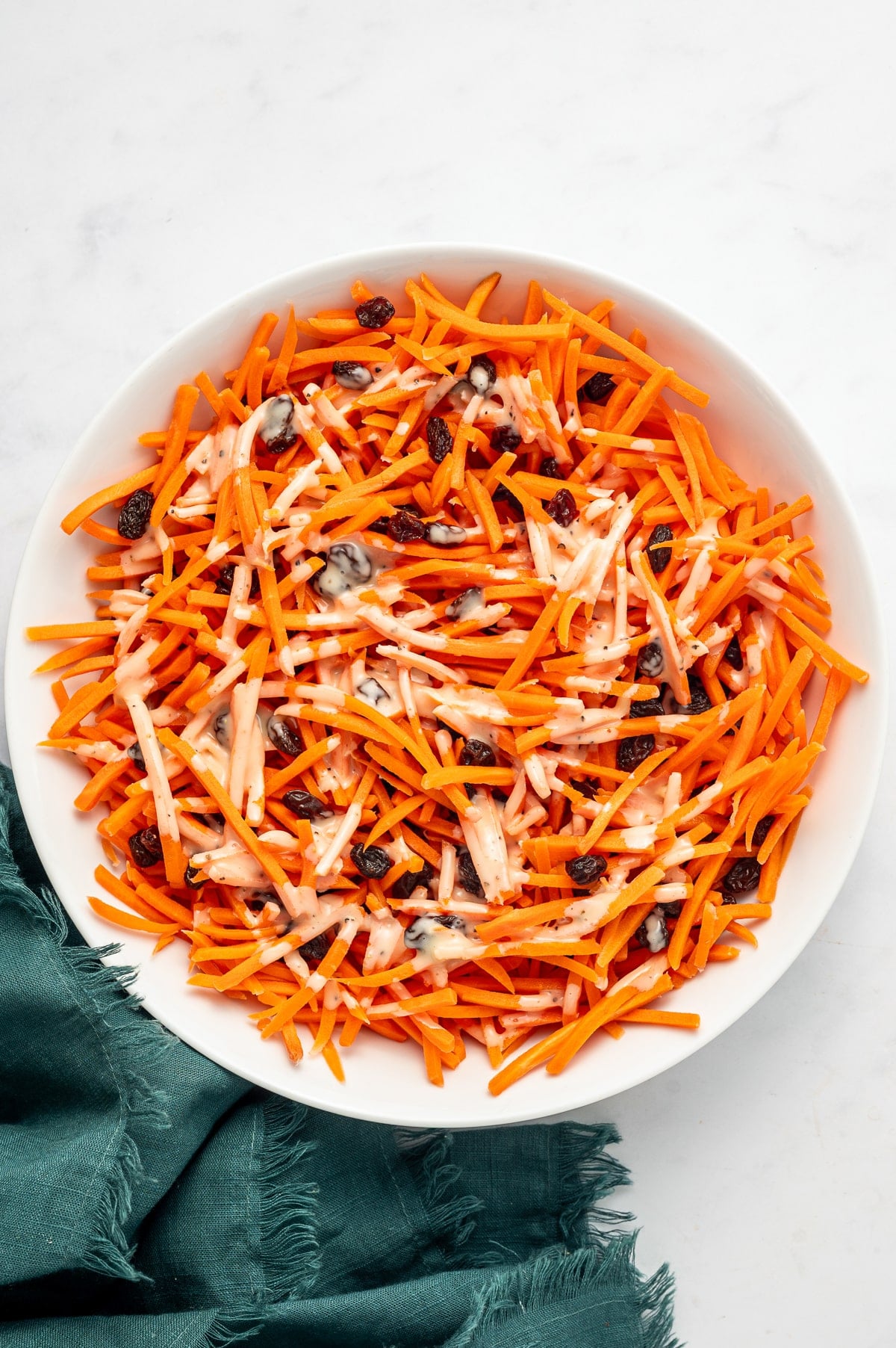 Shredded carrots and raisins in a white oval bowl with creamy honey-lemon mayonnaise dressing drizzled over the top on a white marble surface with a green cloth napkin.