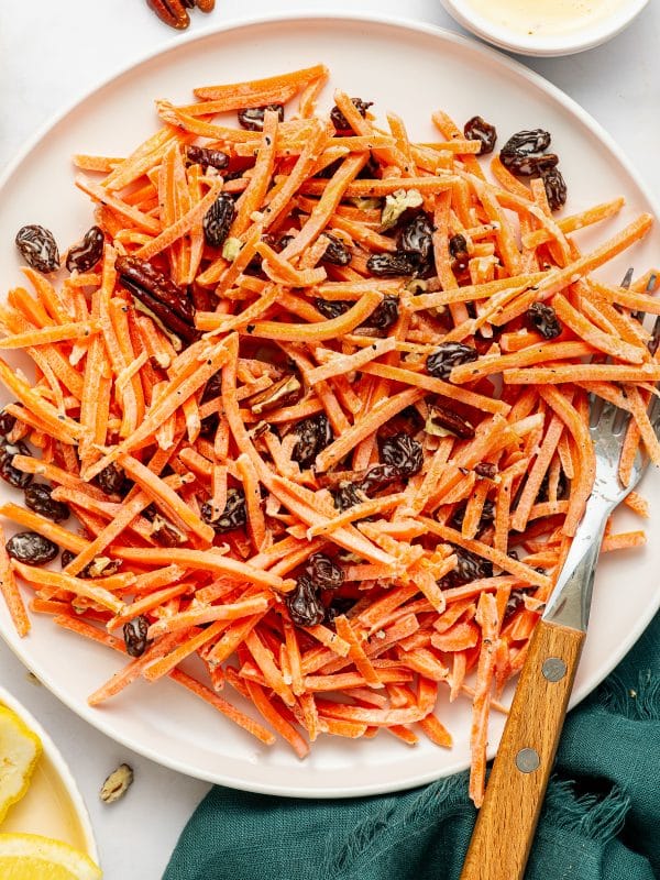 Carrot raisin salad on a white oval plate with shredded carrots, raisins, chopped pecans, creamy dressing, black pepper, and a wooden-handled serving fork.