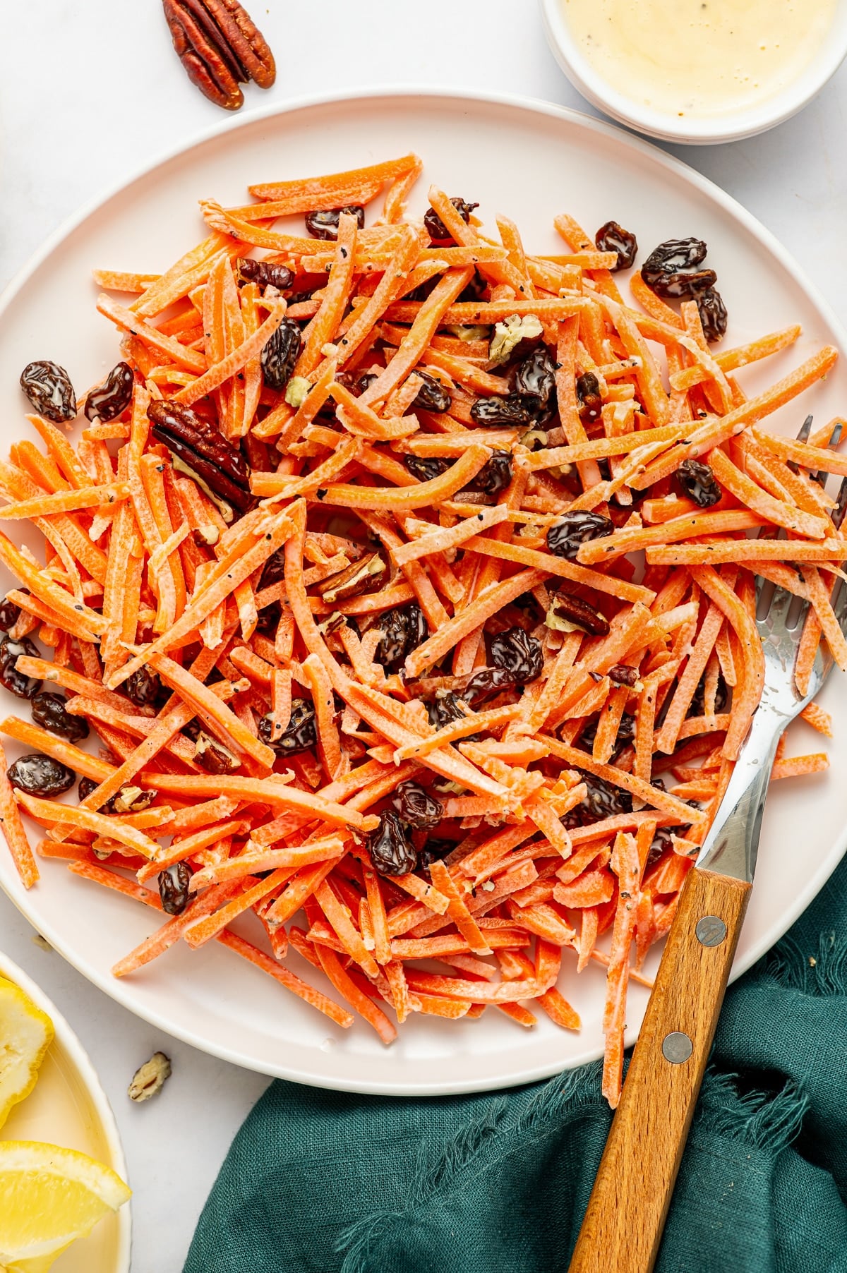 Carrot raisin salad on a white oval plate with shredded carrots, raisins, chopped pecans, creamy dressing, black pepper, and a wooden-handled serving fork.